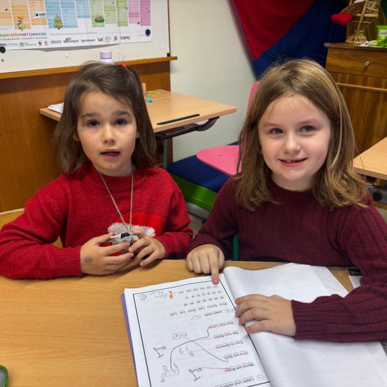 Zwei junge Mädchen sitzen an einem Schreibtisch in einem Klassenzimmer. Sie arbeiten an einem Arbeitsbuch mit Zeichnungen und Diagrammen. Das Mädchen auf der linken Seite hält ein kleines Spielzeug.