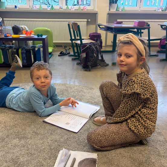 Zwei Kinder sitzen auf dem Teppich in einem Klassenzimmer. Ein Kind liegt mit einem Buch auf dem Boden, das andere sitzt mit einem Stift und einem Notizbuch. Dahinter befinden sich Stühle, Tische und ein Regal mit verschiedenen Gegenständen.
