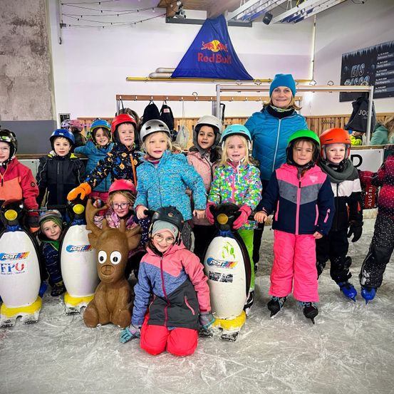 Eine Gruppe von Kindern und einer erwachsenen Frau posiert für ein Foto auf einer Eisbahn. Sie tragen alle Winterkleidung und Helme. Vor ihnen stehen Pinguinstatuen und eine Rentierstatue.