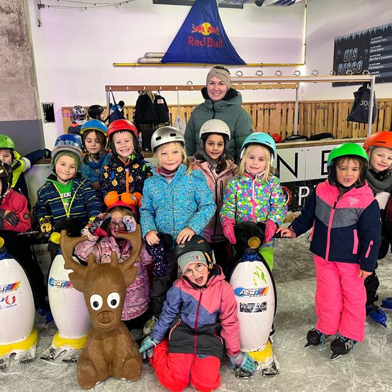 Eine Gruppe von Kindern, alle in Winterkleidung und Helmen, posiert für ein Foto mit einer Pinguin-Statue und einer Rentier-Statue auf einer Eisbahn. Ein Mann steht lächelnd hinter ihnen.
