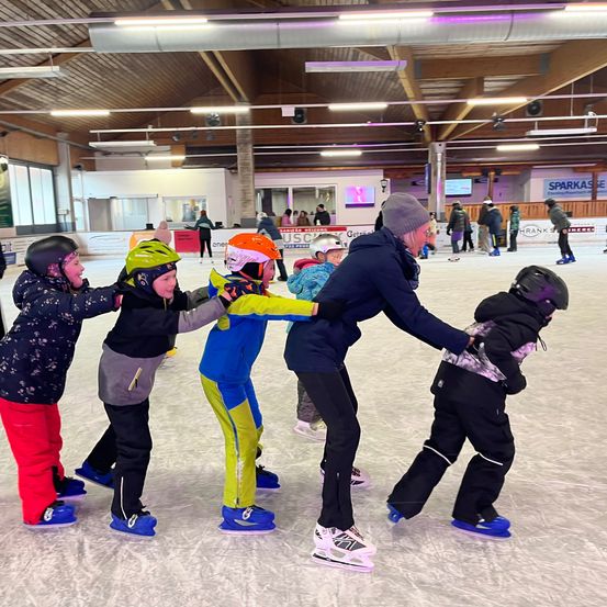 In einer überdachten Eisbahn halten eine Gruppe von Kindern mit Helmen und Eisschuhen Händchen, während eine Frau in einer Jacke ihnen hilft. Die Eisbahn hat Holzdecken mit Lampen und ein Schild an der Wand.