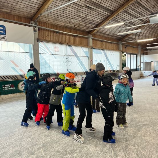 Eine Gruppe von Kindern, die mit Erwachsenen auf einer Eisbahn in einer Indoor-Eisbahn Schlittschuh laufen, tragen Helme und Schlittschuhe, eine Person im Hintergrund.