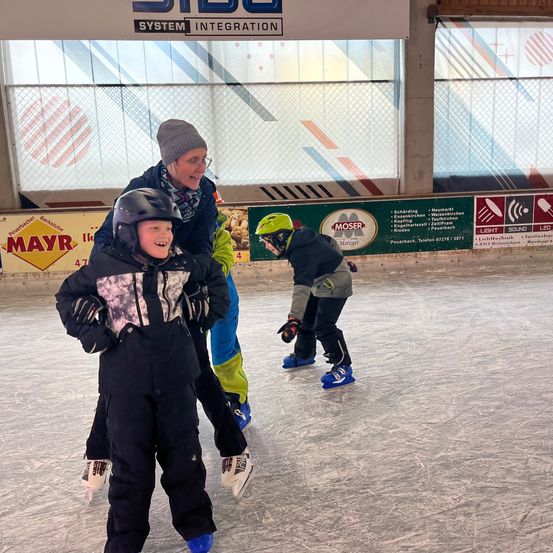 Drei Menschen auf einer Eisbahn im Innenbereich, zwei Kinder und ein Erwachsener. Ein Kind trägt einen Helm und lächelt. Dahinter hält eine Frau mit Brille ein Kind. Ein Schild an der Wand steht 'MOSER'.