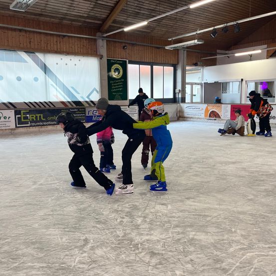 Eine Eislaufbahn mit Holzwänden und Glasfenstern. Mehrere Personen sind beim Eislaufen, darunter zwei Erwachsene, die Kindern beim Schlittschuhlaufen helfen. Ein Banner mit Text ist an der Wand angebracht.