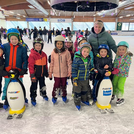 Eine Gruppe von Kindern mit pinguinförmigen Eislaufschuhen, die Winterkleidung tragen, posiert für ein Foto auf einer Indoor-Eisbahn. Eine erwachsene Frau steht hinter ihnen.