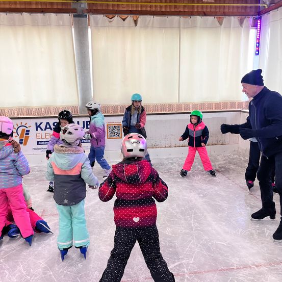 Eine Gruppe von Kindern in Schlittschuhen und Helmen fährt auf einer überdachten Eisbahn. Ein erwachsener Ausbilder steht in der Nähe und beaufsichtigt sie.