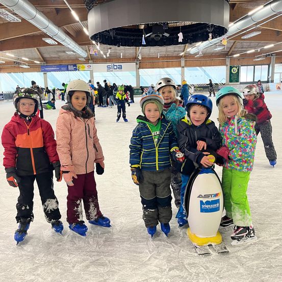 Eine Gruppe von Kindern auf einer Eisbahn, in Winterkleidung und Helmen, stehen lächelnd zusammen. Hinter ihnen ist ein Pinguin-Maskottchen zu sehen. Die Eisbahn hat ein Innensetting mit Lampen an der Decke.