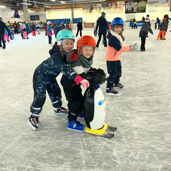 Zwei Kinder mit Helmen skaten auf einer Eisbahn mit anderen. Ein Kind hält ein pinguinförmiges Objekt. Die Halle ist mit weißer Decke und Werbung an den Wänden ausgestattet.
