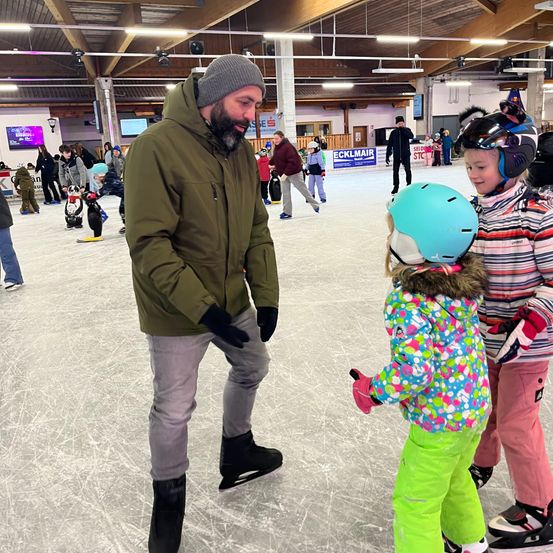 Ein Mann hilft zwei jungen Mädchen auf einer Eisbahn. Ein Mädchen trägt einen Helm und Handschuhe. Ein anderes Kind spielt im Hintergrund.