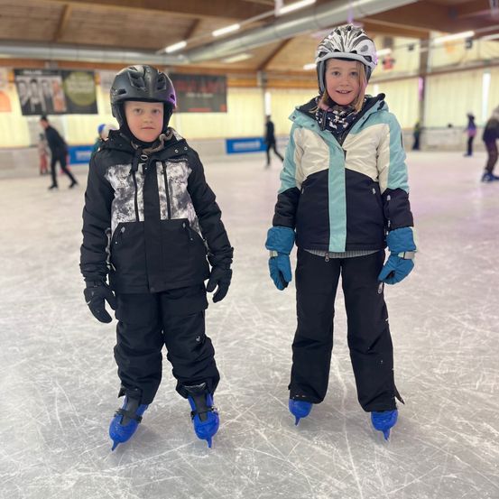 Zwei Kinder stehen auf einer Eisbahn, tragen Winterjacken, Helme und Schlittschuhe. Sie lächeln und schauen nach vorne. Die Eisbahn hat weiße Wände und eine Holzdecke mit Lichtern.