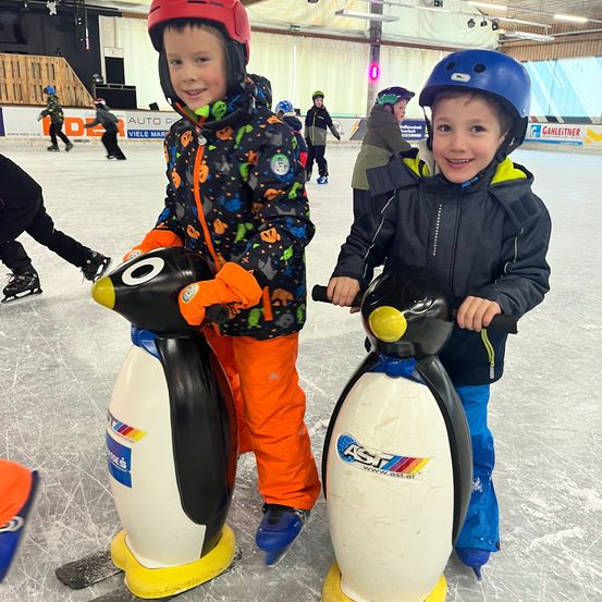 Zwei Jungen in Winterkleidung stehen auf einem Eislaufplatz auf pinguinförmigen Rollern, lächeln und halten die Griffe fest. Hinter ihnen skaten mehrere andere Personen, und die Halle ist mit Werbung dekoriert.