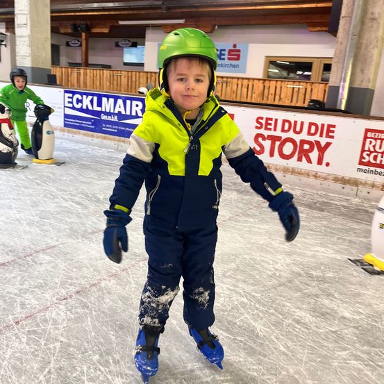 Ein junger Junge mit einem grünen Helm und einem bunten Mantel skat auf einer Eisbahn in einer Halle. Hinter ihm steht ein Pinguin-Maskottchen auf dem Eis. An den Wänden sind Banner mit Text angebracht.
