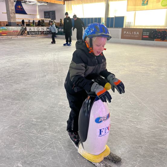 Ein junger Junge mit blauem Helm und Handschuhen reitet auf einem pinguinförmigen Spielzeug auf einer überdachten Eisbahn. Hinter ihm skaten zwei Personen. Die Bahn hat Schilder und Banner an den Wänden.