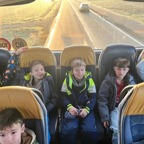 Fünf Jungen sitzen in einem Bus mit Straßenblick, zwei Autos dahinter. Ein Straßenschild zeigt die Geschwindigkeitsbegrenzung 80 an.