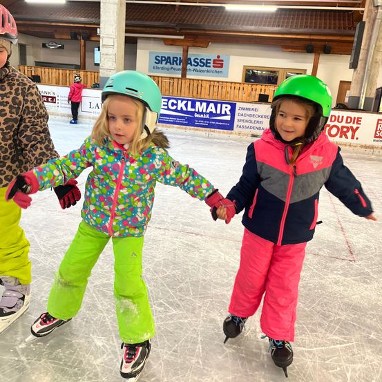 Zwei kleine Mädchen in Helmen und Schlittschuhen halten sich an den Händen, während sie in einer überdachten Eisbahn Schlittschuh laufen. Hinter ihnen beobachten ein Mensch in einem Leoparden-Jacke und ein Kind in einer pinken Jacke.