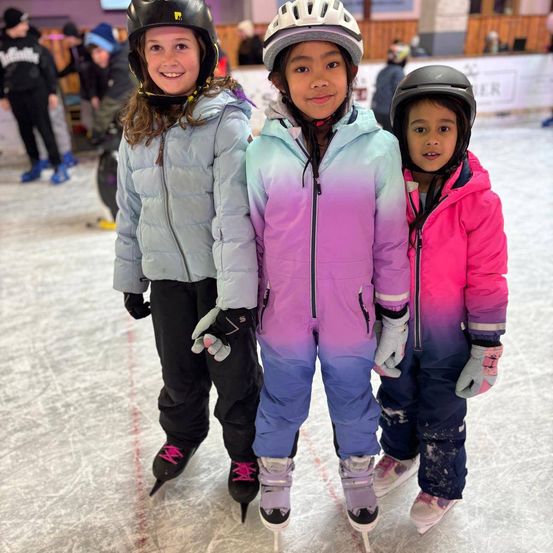 Drei Kinder stehen auf einer Eisbahn und tragen Winterkleidung und Helme. Das Mädchen auf der linken Seite trägt eine graue Jacke, die in der Mitte eine lila-pinke Kombination und das Mädchen auf der rechten Seite eine rosa Jacke.