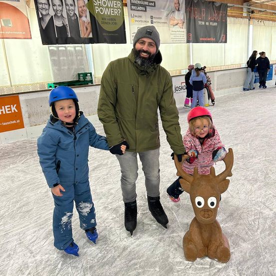 Ein Mann und zwei Kinder posieren mit einer Rentierdekoration auf einer überdachten Eisbahn.