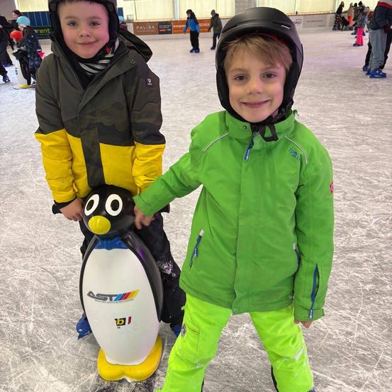 Zwei Jungen in Winterkleidung stehen auf einer Eisbahn. Ein Junge hält einen pinguinförmigen Roller. Hinter ihnen schieben sich Menschen auf Schlittschuhen.