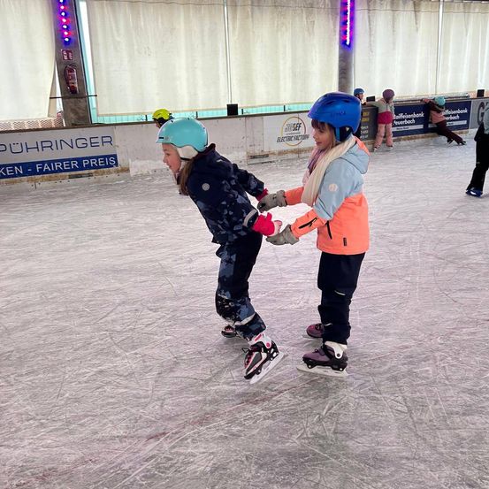 Zwei Kinder in Helmen und Handschuhen skaten gemeinsam auf einer überdachten Eisbahn. Das Mädchen vorne trägt eine blaue Jacke, während das dahinterliegende ein orangefarbenes Jackett trägt. Die Eisbahn hat eine weiße Barriere mit einem Schild, auf dem 'Puehringer' steht.