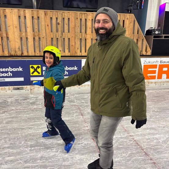 Ein Mann und ein Kind skaten auf einer Eisbahn. Das Kind trägt einen gelben Helm und blaue Handschuhe. Der Mann trägt eine grüne Jacke und einen Bart. Hinter ihnen steht eine Holzbarriere.