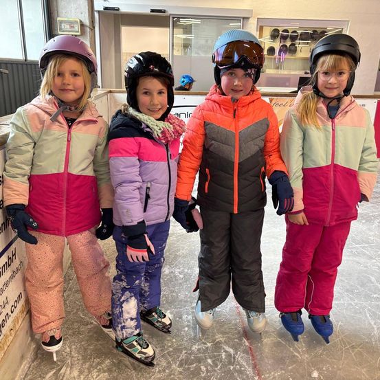 Vier junge Mädchen stehen zusammen auf einer Eisbahn, alle tragen Winterjacken, Hosen und Helme. Jedes trägt Eisschuhe. Sie scheinen für ein Foto zu lächeln.