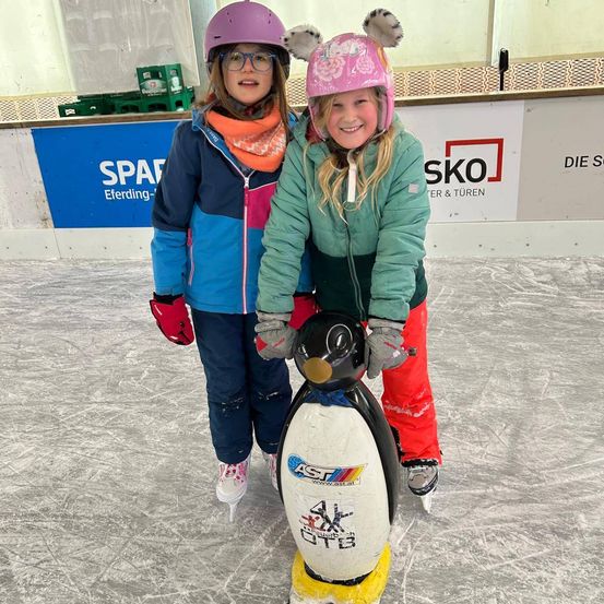 Zwei junge Mädchen in Winterkleidung eislaufen in einer überdachten Eisbahn. Eine fährt einen pinguinförmigen Roller, während die andere neben ihr lächelt.