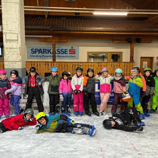 Eine Gruppe von Kindern in Winterkleidung posiert für ein Foto auf einer überdachten Eisbahn, wobei zwei Kinder auf dem Eis liegen.
