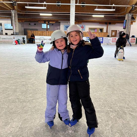 Zwei junge Mädchen in Winterkleidung posieren für ein Foto auf einer Indoor-Eisbahn. Ein Mädchen macht ein Friedenszeichen. Im Hintergrund skaten andere Leute, während einige sitzen.