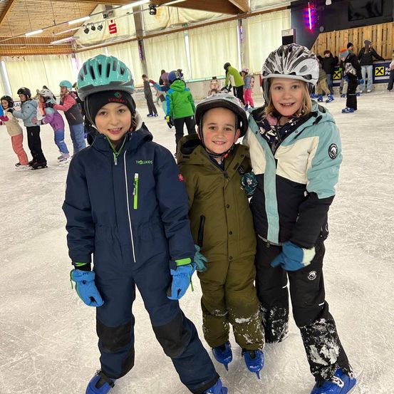 Drei Kinder stehen gemeinsam auf einer überdachten Eisbahn, lächeln und posieren für ein Foto. Sie tragen Winterkleidung und Helme. Im Hintergrund skaten andere Kinder und Menschen stehen oder sitzen.