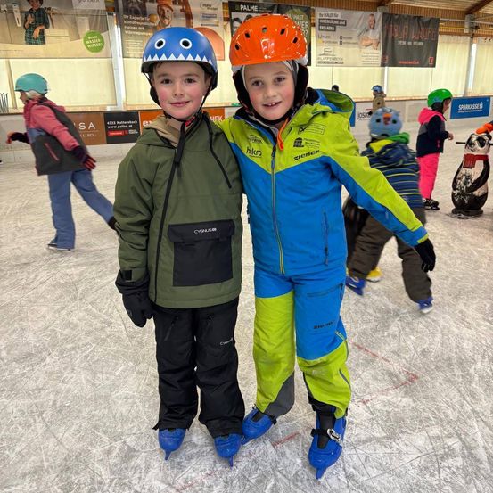 Zwei junge Jungen, einer in einer grünen Jacke und der andere in einem blau-grünen Outfit, stehen auf einer Eisbahn. Beide tragen Helme und Schlittschuhe. Hinter ihnen sind auch andere Kinder beim Schlittschuhlaufen.