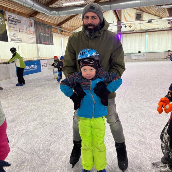 Ein Mann in Mütze und Jacke hält ein Kind in Helm und Handschuhen, das auf einer Eisbahn steht, mit anderen Leuten und Werbung an den Wänden.