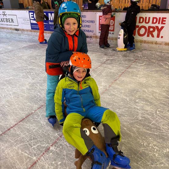 Zwei Kinder mit Eislaufausrüstung auf einer Eisbahn. Eines steht, das andere sitzt auf einem pinguinförmigen Schlitten.