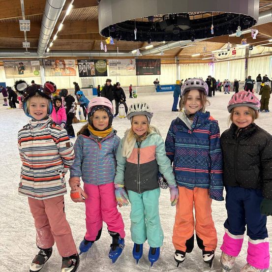 Fünf Kinder, alle mit Helmen und Schlittschuhen bekleidet, stehen zusammen auf einer überdachten Eisbahn. Sie lächeln und posieren für ein Foto.