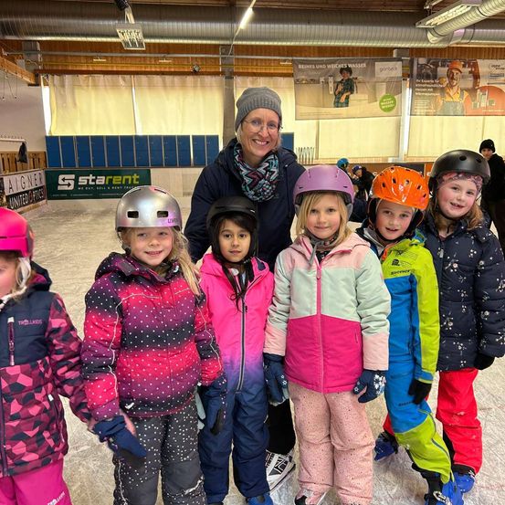 Eine Gruppe von Kindern in Winterkleidung steht auf einer Eisbahn mit einer lächelnden Frau. Sie tragen alle Helme und einige haben Skier dabei. Ein Banner für Starent ist hinter ihnen zu sehen.