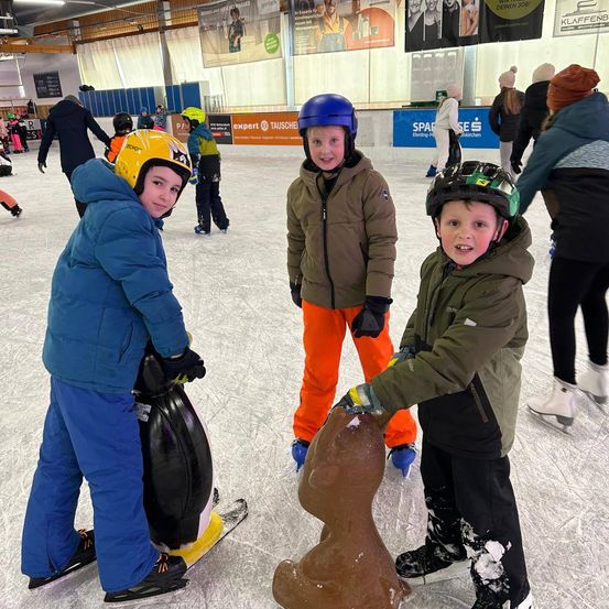 Drei junge Jungen sind Eislaufen in einer überdachten Eishalle. Sie tragen Helme und Handschuhe. Ein Junge hält ein pinguinförmiges Objekt, ein anderer ein Snowboard. Hinter ihnen sind weitere Skater und Zuschauer anwesend, mit Werbung an den Wänden.