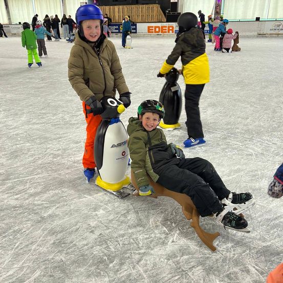 Zwei Kinder sind auf einer Eisbahn. Eins sitzt auf einem pinguinförmigen Schlitten, und das andere steht neben ihm. Andere Kinder skaten umher.