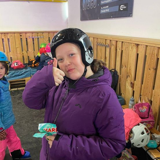 Ein junges Mädchen in einem lila Mantel und Helm zeigt mit einem Daumen nach oben, während es einen Snack hält. Hinter ihr sitzen andere mit Helmen und Ausrüstung an hölzernen Wänden.