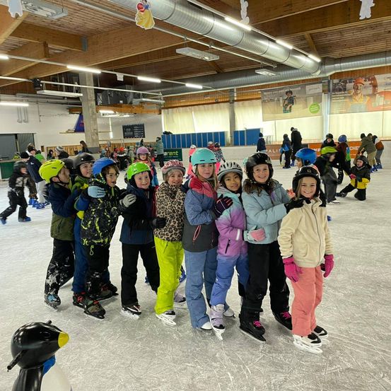 Eine Gruppe von Kindern in Winterkleidung und Helmen, die lächelnd und für ein Foto auf einer Eisbahn in einer Halle posieren.