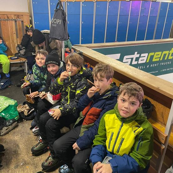 Eine Gruppe junger Jungen sitzt auf einer Bank in einer Umkleidekabine, isst und unterhält sich. Ein Junge trägt einen schwarzen und gelben Mantel. Ein Schließfach mit einer Tasche hängt über ihnen.