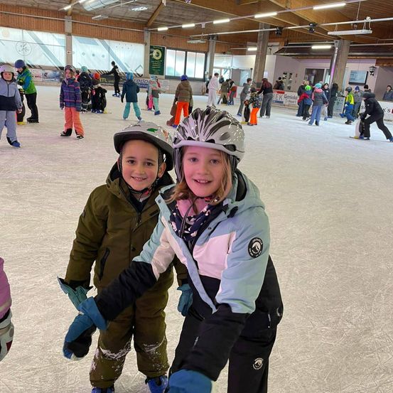 Zwei Kinder mit Helmen skaten auf einer Eisbahn, umgeben von einer Menge Leute.