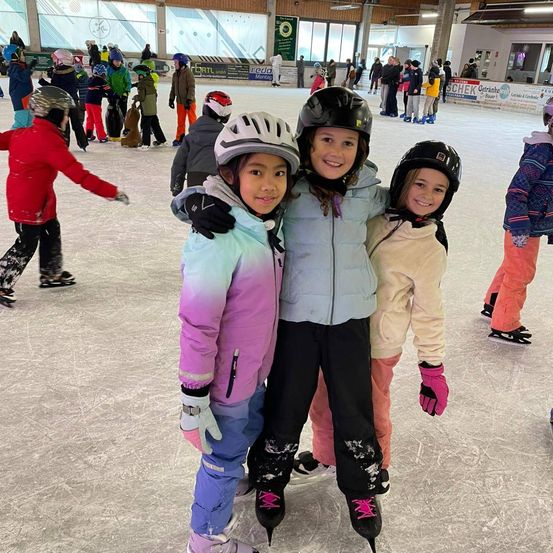 Drei Mädchen in Winterkleidung und Helmen posieren für ein Foto auf einer Indoor-Eisbahn, mit anderen Skatern im Hintergrund.