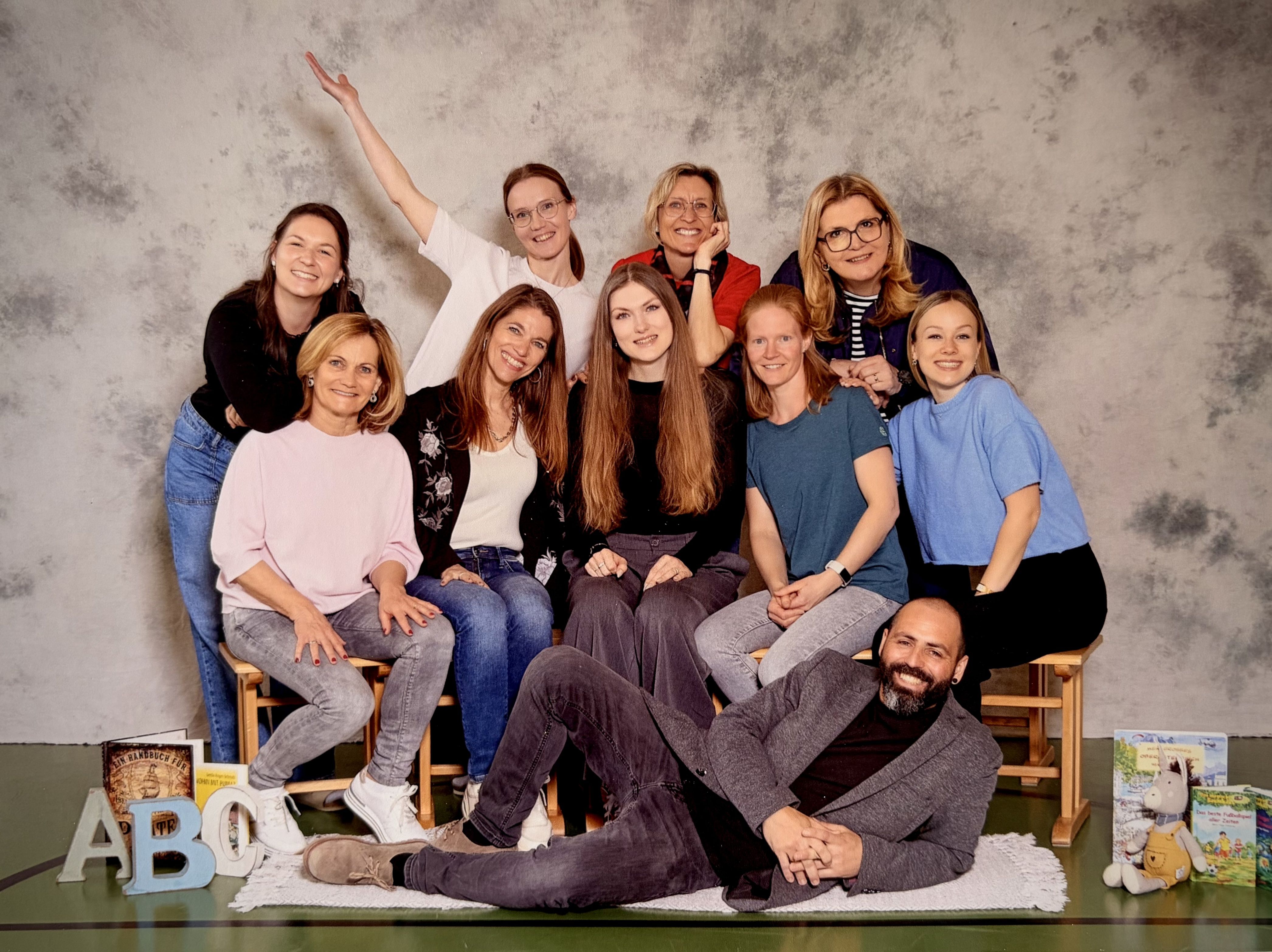 A group of women and a man are posing for a photo with smiles on their faces. They are sitting on wooden chairs and standing against a textured wall.