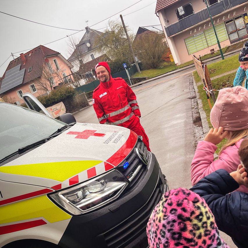 Eine Person in einem roten Anzug steht neben einem Krankenwagen mit Kindern in der Nähe. Die Szene spielt sich auf einer Straße mit Häusern und Solarpanelen ab.