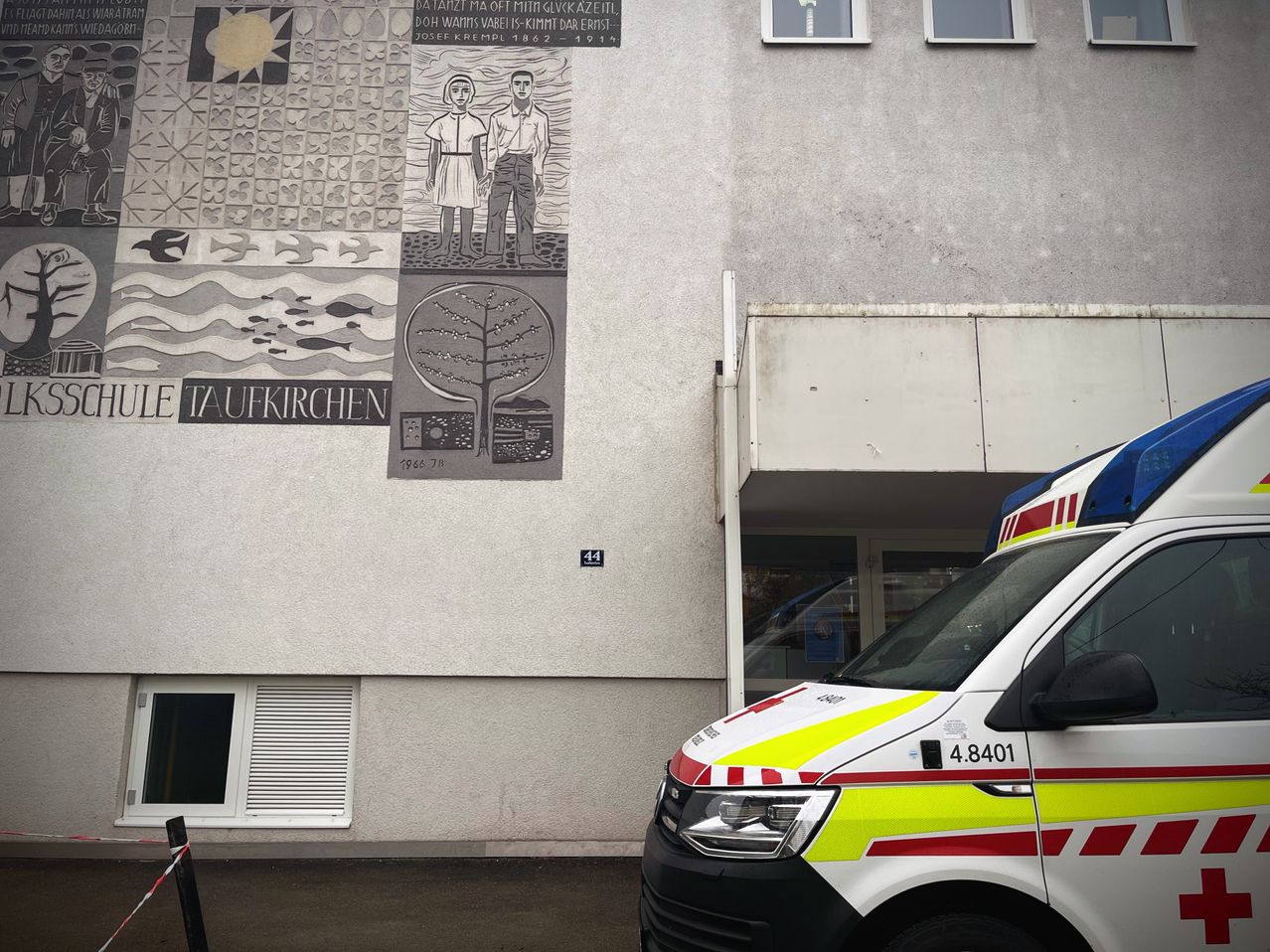 Ein Krankenwagen steht vor einem Gebäude mit einem Wandbild von zwei Personen und einem Baum. Das Gebäude hat Fenster und eine Tür.