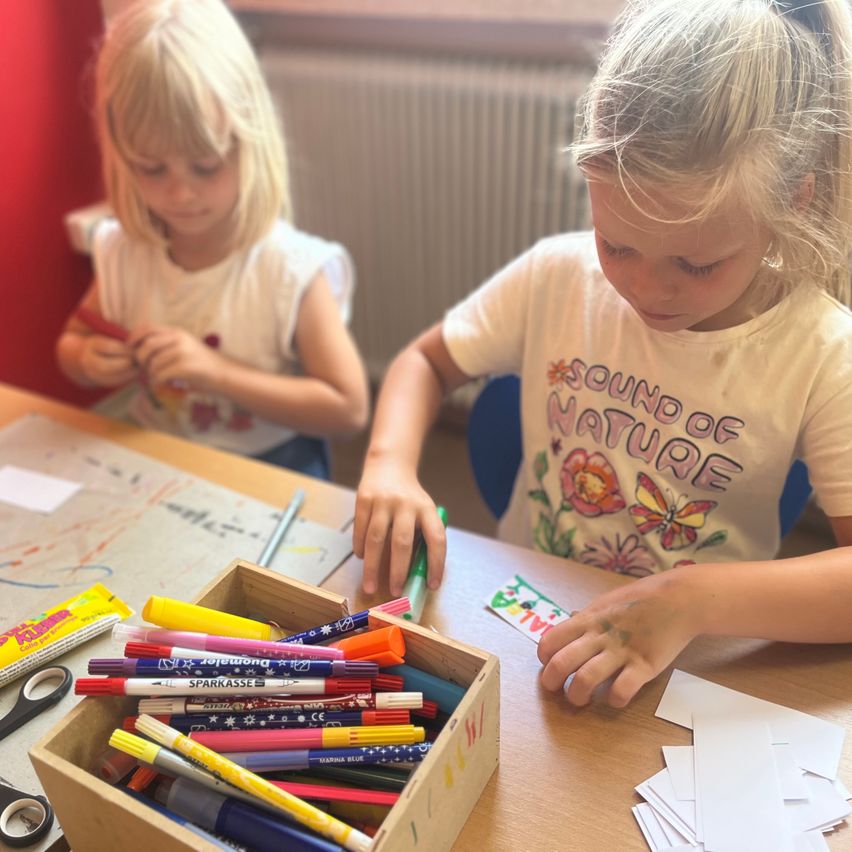 Two girls sit at a table, engaged in a coloring activity. One is coloring a paper while the other sits nearby. A box full of crayons and markers sits on the table.