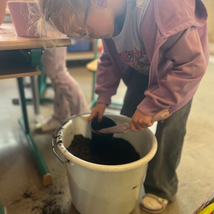 A child pours soil into a bucket, wearing a pink jacket and glasses, inside a classroom.