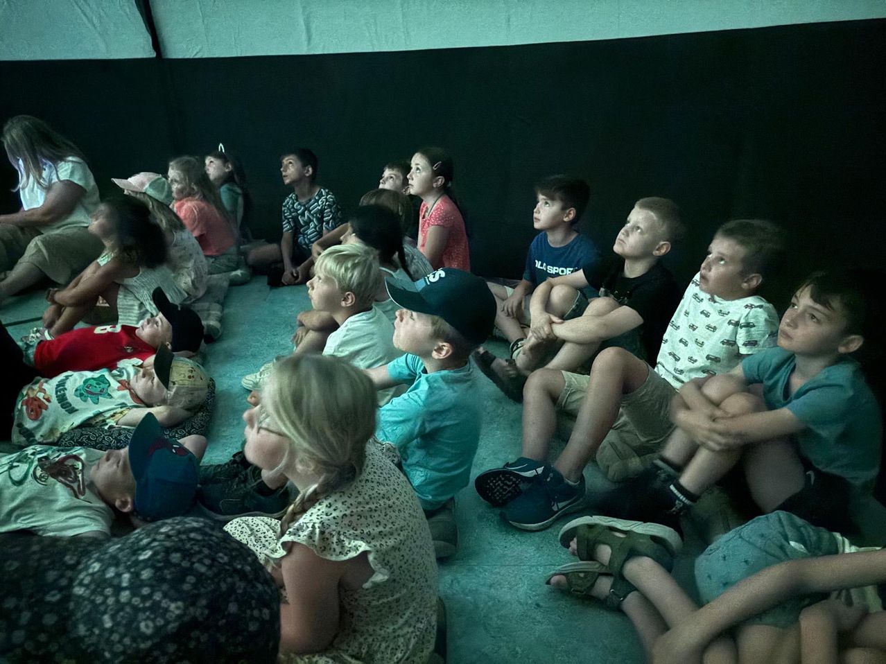 Eine Gruppe von Kindern sitzt auf dem Boden, einige mit Mützen, einige mit den Händen am Kinn, in Richtung eines unsichtbaren Bildschirms.