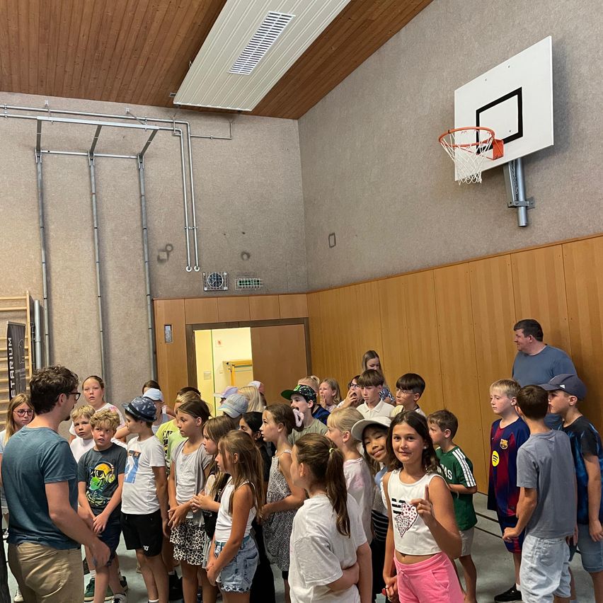 Eine Gruppe von Kindern steht in einer Turnhalle, einige tragen Mützen. Ein Mann in einem blauen T-Shirt spricht zu ihnen. Ein Basketballkorb ist an einer Wand montiert, und Holzpaneele säumen die Wände.