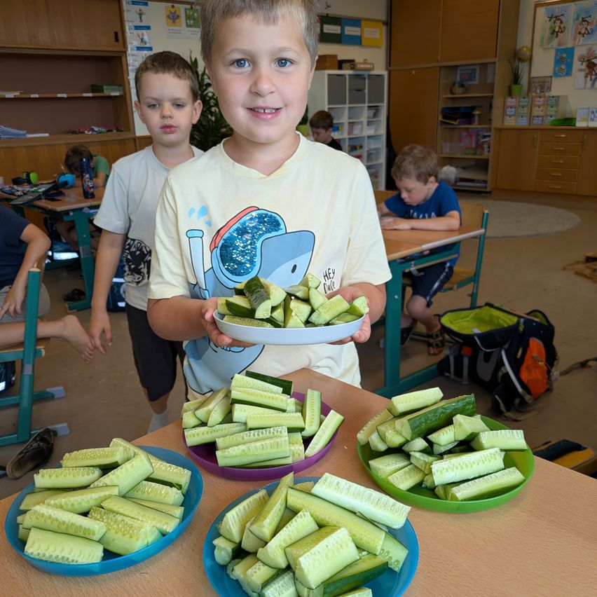 Ein junger Junge mit einem lächelnden Gesicht hält einen Teller mit geschnittenen Gurken in einem Klassenzimmer. Hinter ihm sitzen zwei andere Jungen an Schreibtischen und einige Kinder stehen im Hintergrund.