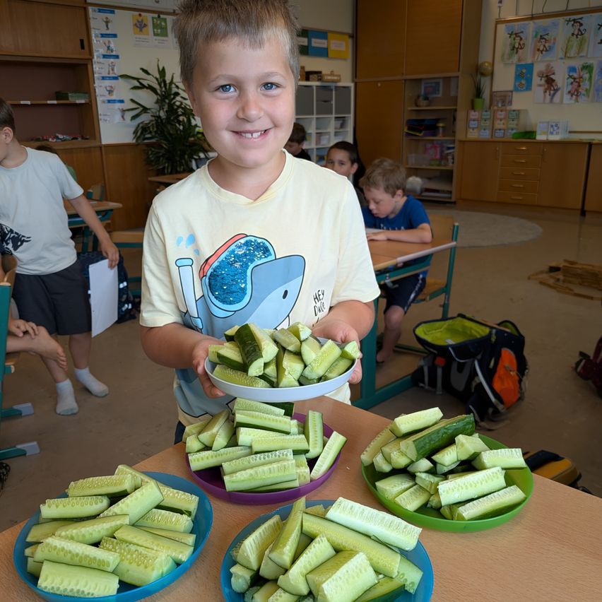 Ein Junge hält eine Platte mit Gurkenscheiben und lächelt im Klassenzimmer. Andere Kinder und Erwachsene sind in der Nähe.